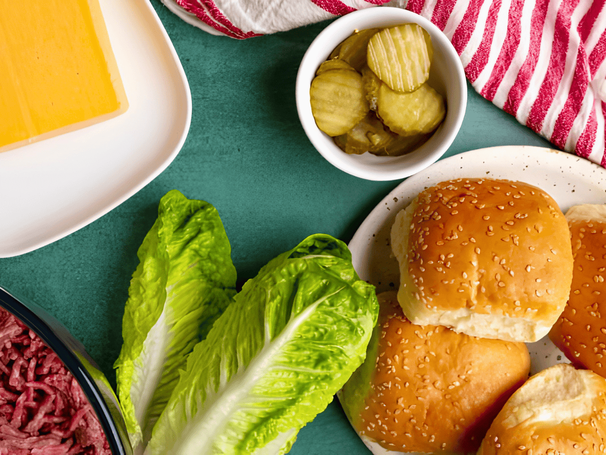 bread buns on a plate with lettuce leaves, ground beef, cheese, and pickles