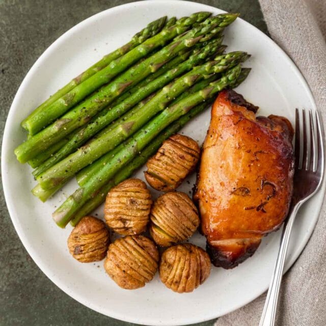 Slow Cooker Honey Lime Chicken thighs plated with mini hasselback potatoes and asparagus with fork on plate, 1x1