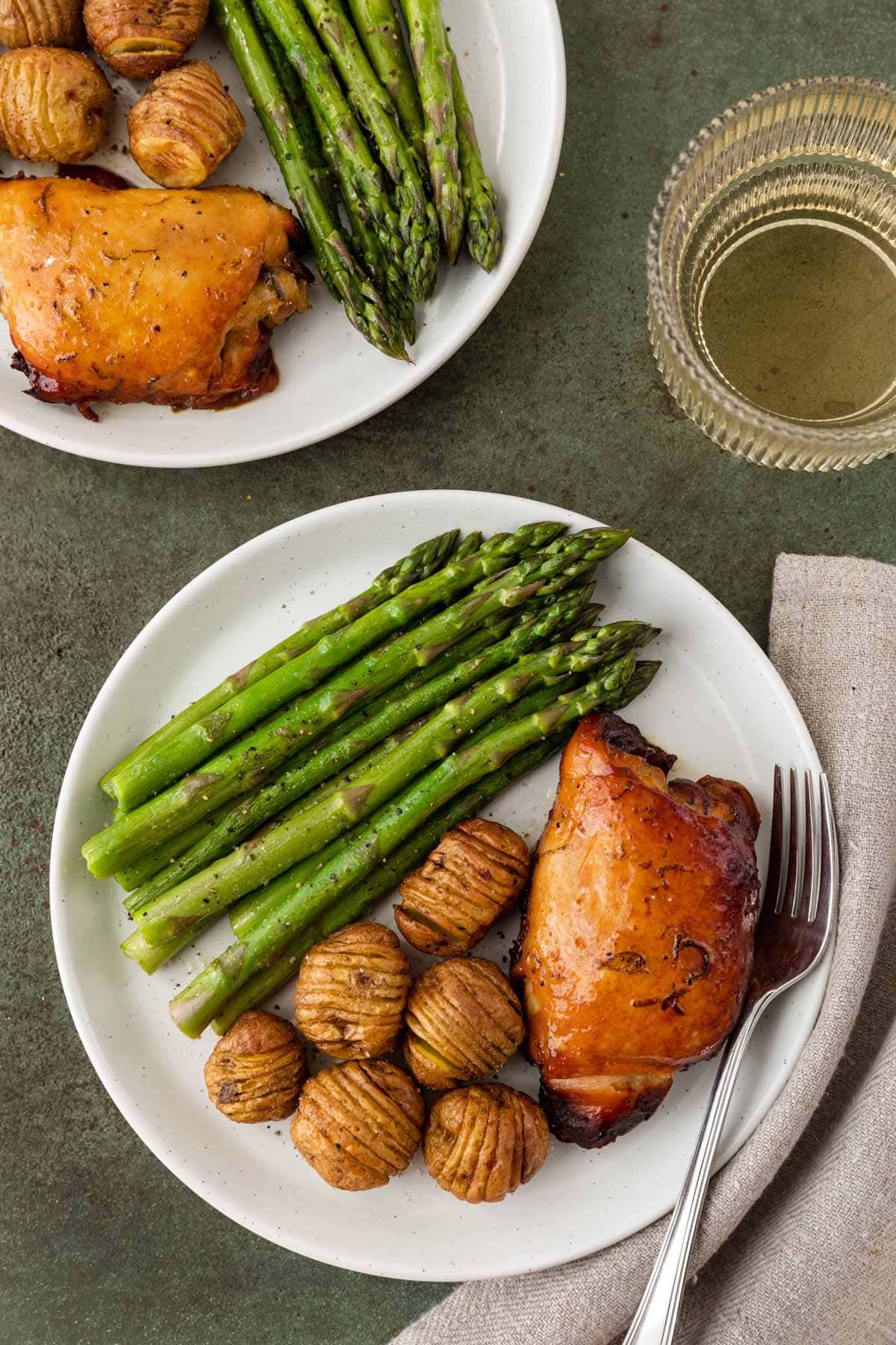Slow Cooker Honey Lime Chicken thighs on two plates with mini hasselback potatoes and asparagus with fork on plate, and glass of wine, top down view