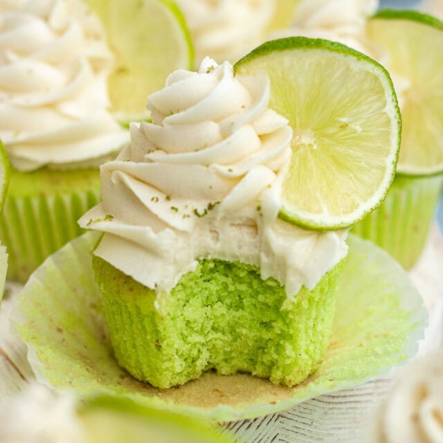 Key Lime Cupcakes frosted cupcakes with lime wheel and lime zest, front cupcake missing bite and wrapper peeled down, 1x1