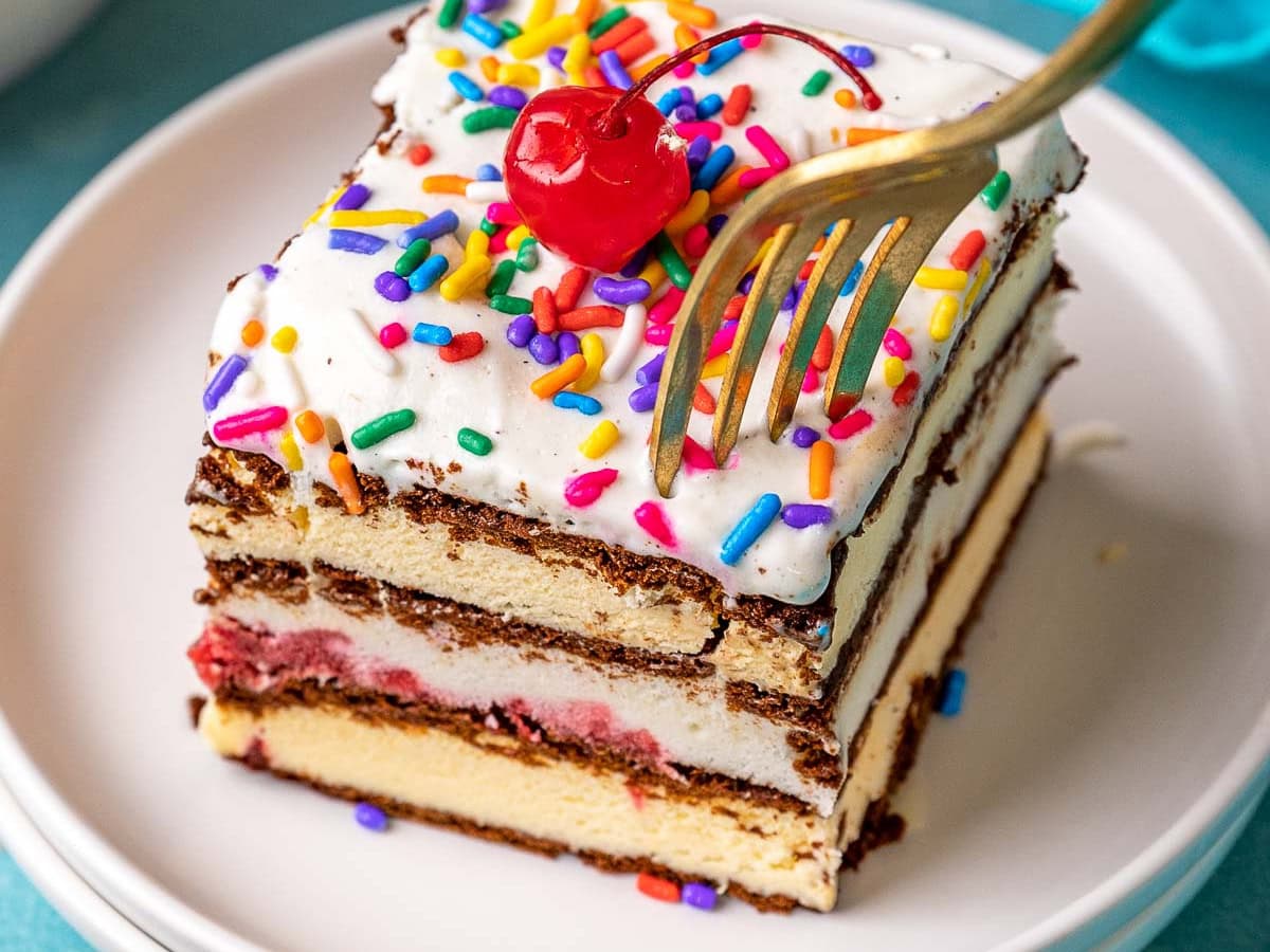 Ice Cream Sandwich Cake slice on plate topped with maraschino cherry. Fork cutting into corner of slice.