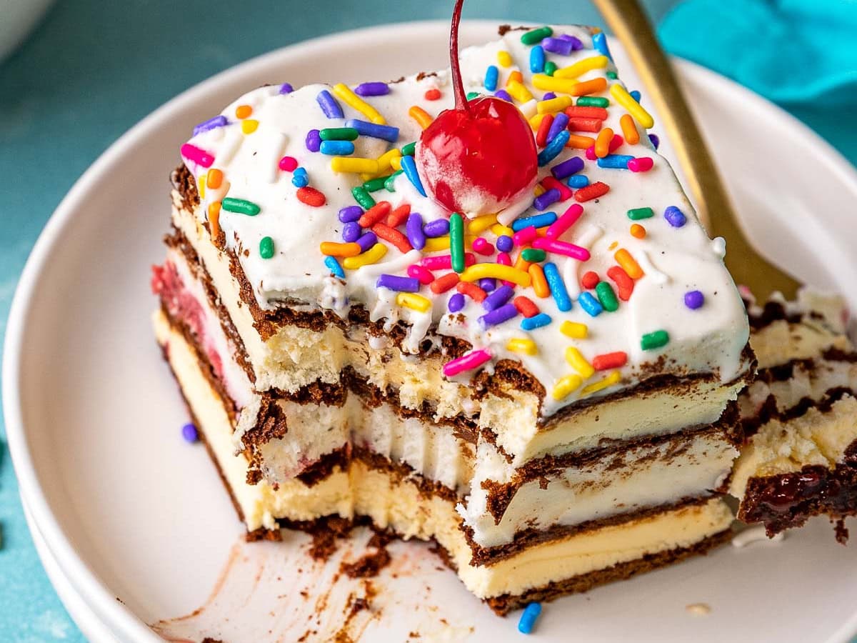 Ice Cream Sandwich Cake slice on plate topped with maraschino cherry. Corner of cake missing. Fork next to slice on plate.