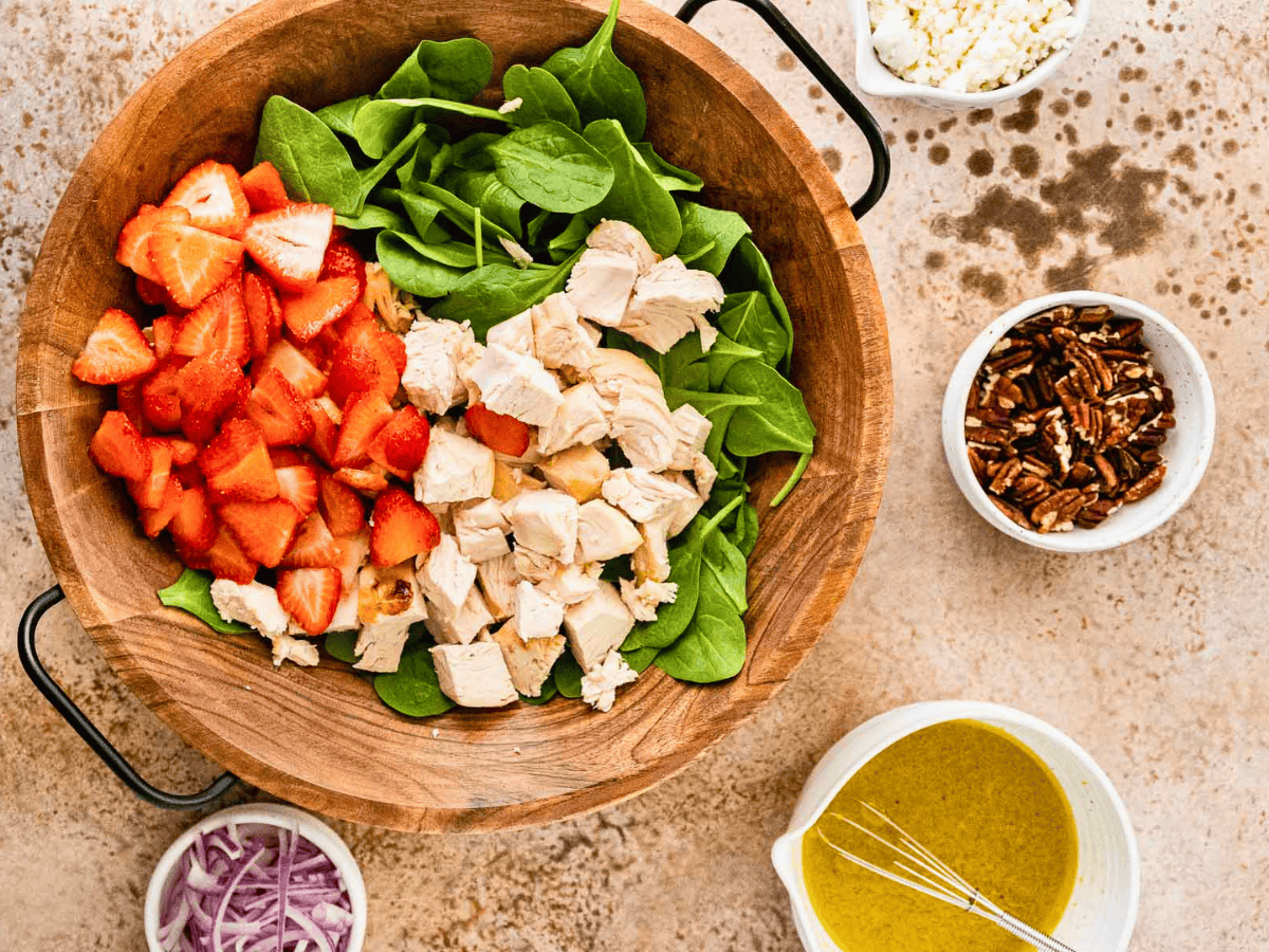 chicken, berries, and spinach in a wooden bowl with pecans in a seperate bowl with dressing on the side