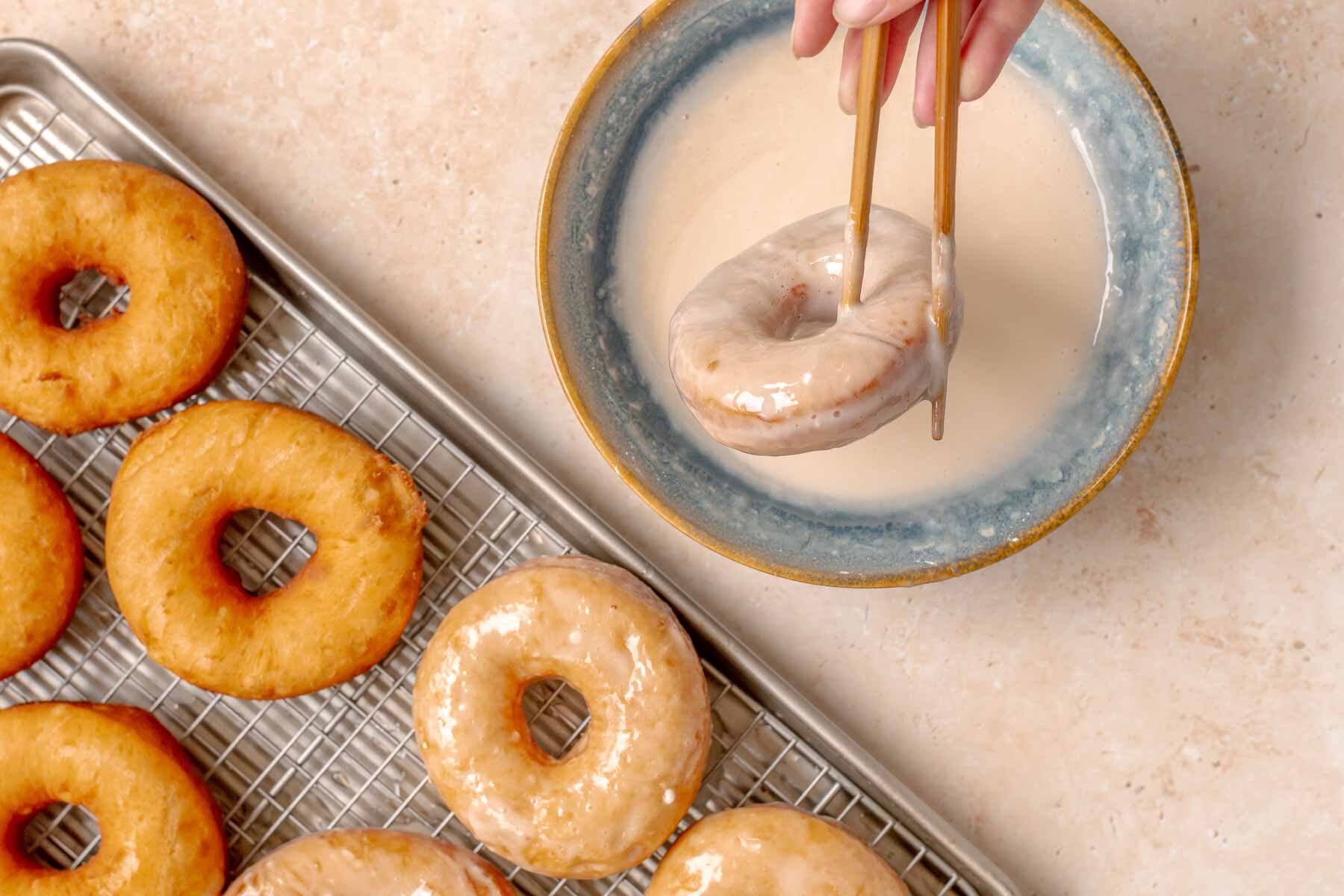 Glazed Potato Donuts dipping donut in glaze with chop sticks, glazed and unglazed donuts on baking sheet.