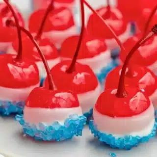 round red fruits with stems dipped in white chocolate and blue sugar side by side on a white plate