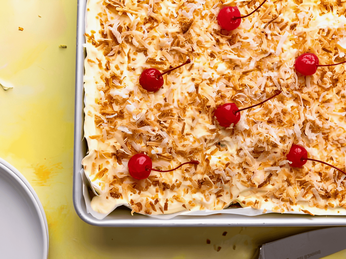 moist tropical cake topped with frosting, coconut, and cherries in a baking pan