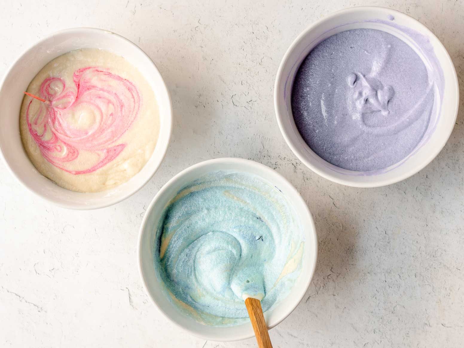 Unicorn Cupcakes pink, blue and purple cake batter in separate bowls. Spatula in blue batter bowl and pink bowl partially mixed.
