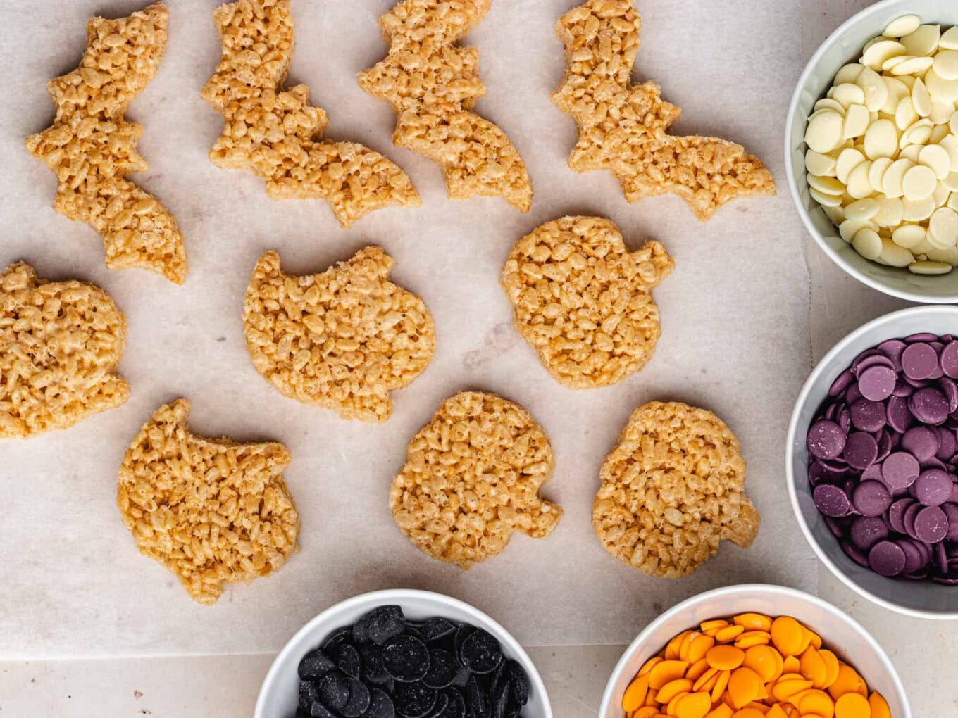 Krispie Treats cut into shapes lined up on counter and bowls of unmelted candy melts framing right side