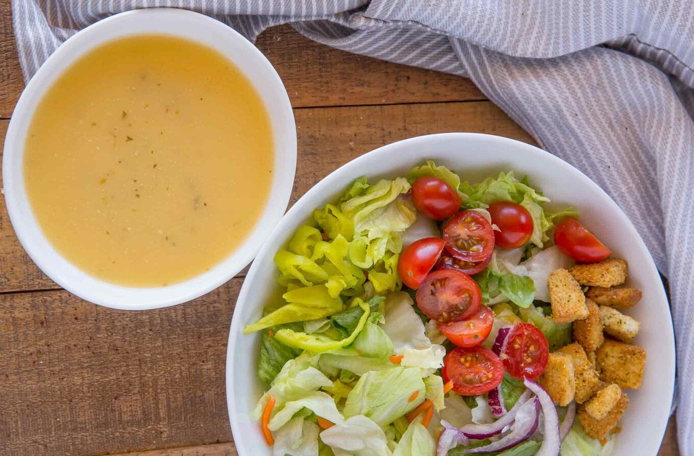 Olive Garden Italian Salad Dressing copycat recipe made with Romano cheese, vinegar, oil and garlic in just seconds tastes exactly like the restaurant version you love. | #olivegarden #copycatrecipes #olivegardendressing #saladdressing #italiandressing #salad #dinnerthendessert Dressing in a small white bowl next to salad in larger bowl.