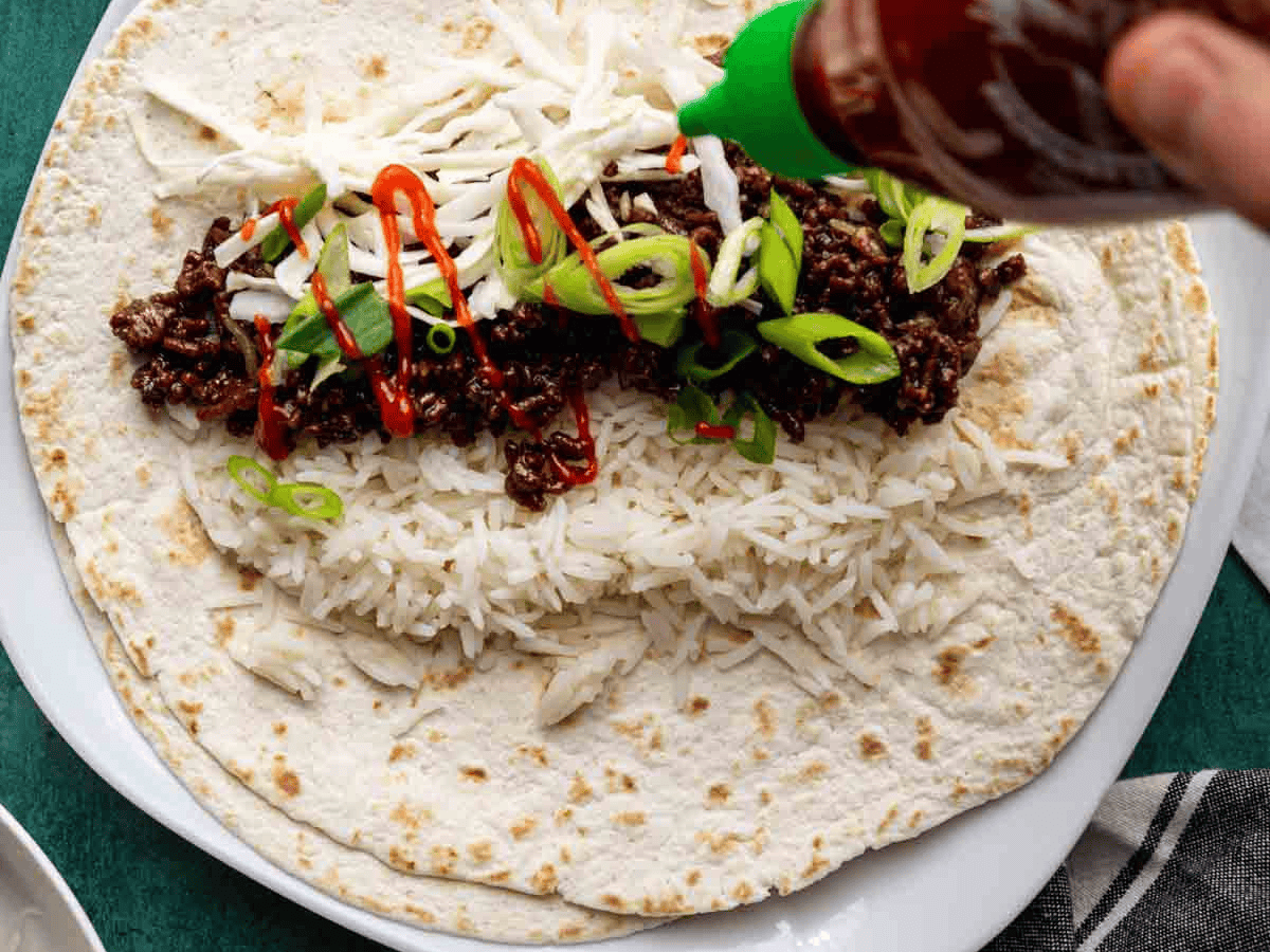 Ingredients being assembled on a tortilla and garnished with a red spicy sauce.