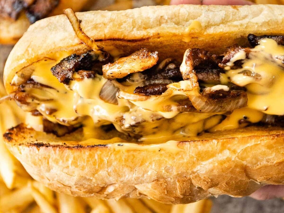 Philly Cheesesteak made in your own kitchen with tender rib eye steak, caramelized onions, and Cheese Whiz, in a buttery hoagie roll. Assembled sandwich held up close.