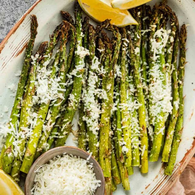Plate of cooked asparagus topped with cheese with lemon wedges