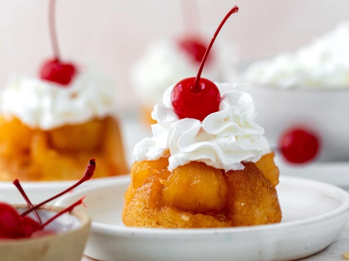 Pineapple Upside Down Cupcakes are a miniature version of the classic dessert with buttery yellow cake, pineapple chunks, and cherries. Cupcakes on serving plates with whipped cream and cherry on top.