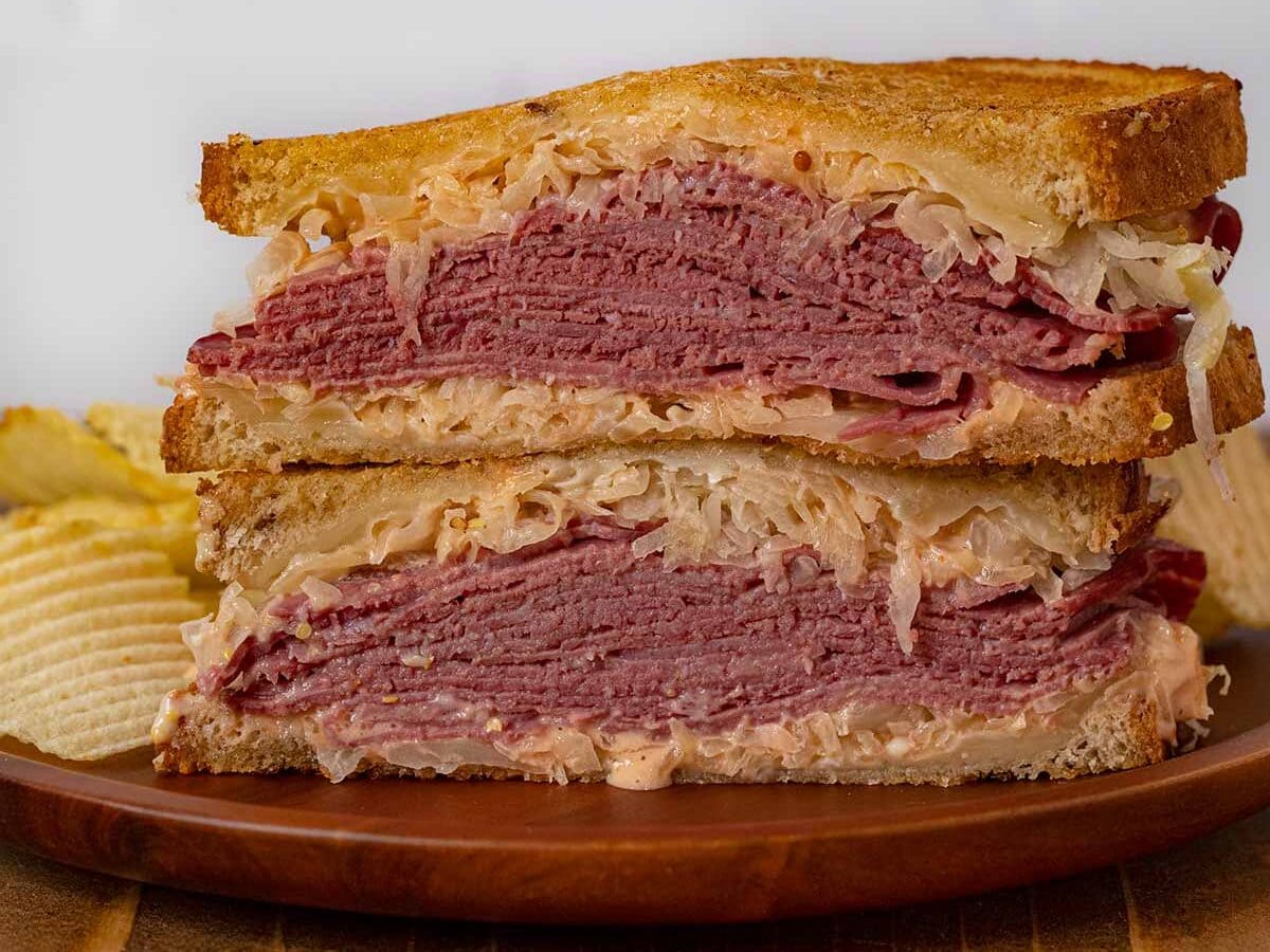 Reuben Sandwiches are the ultimate grilled sandwich, filling and flavorful with corned beef, dressing, Swiss cheese and sauerkraut all on rye bread. #dinner #lunch #sandwich #reuben #cornedbeef #sauerkraut #delisandwich #dinnerthendessert Assembled Sandwich halves in stack on plate with chips