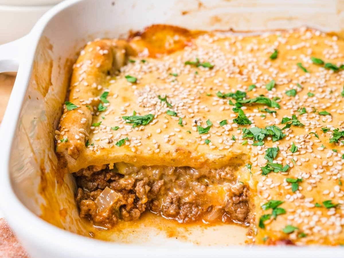 Easy Sloppy Joe Casserole is a tasty quick family friendly dinner. Baked sesame seed bun topping with a cheesy ground beef Sloppy Joes filling from scratch. Side view of baked casserole in dish with slice removed