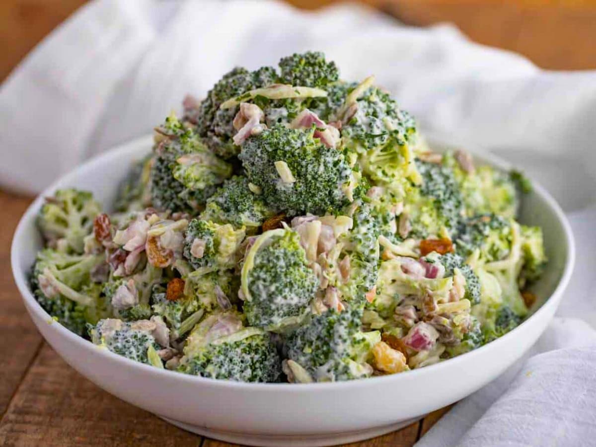 Broccoli Salad is an easy summer side with raisins, bacon and sunflower seeds. A salad can sit without wilting is perfect for outside events! Dressed salad with raisins in a white bowl