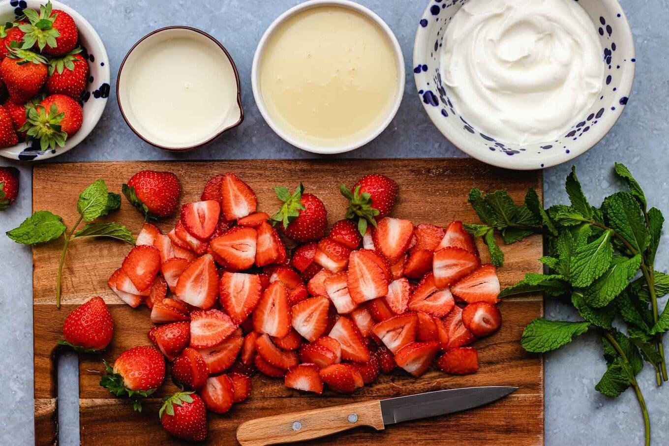 Fresas con Crema, or strawberries with cream, is an easy, creamy, no-bake Mexican dessert ready in 15 minutes with 5 simple ingredients! Halved strawberries on wooden board, remaining ingredients in prep bowls.