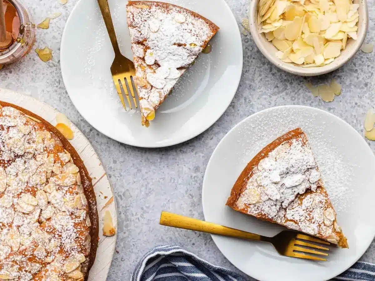 Top view of multiple slices of cake on top of white dishes with forks with powder sugar and almonds on top