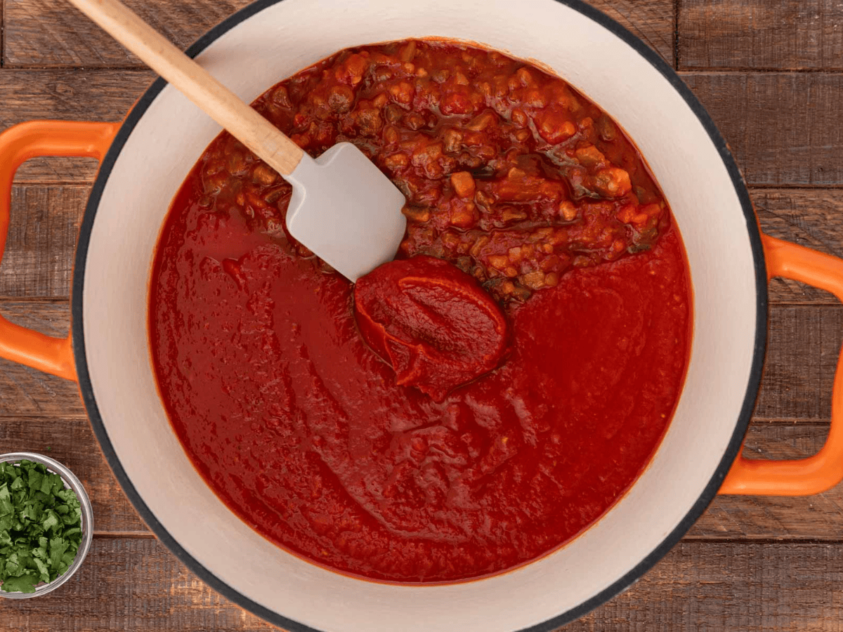Tomato sauce being added to a white pot with cooked meat mixture.