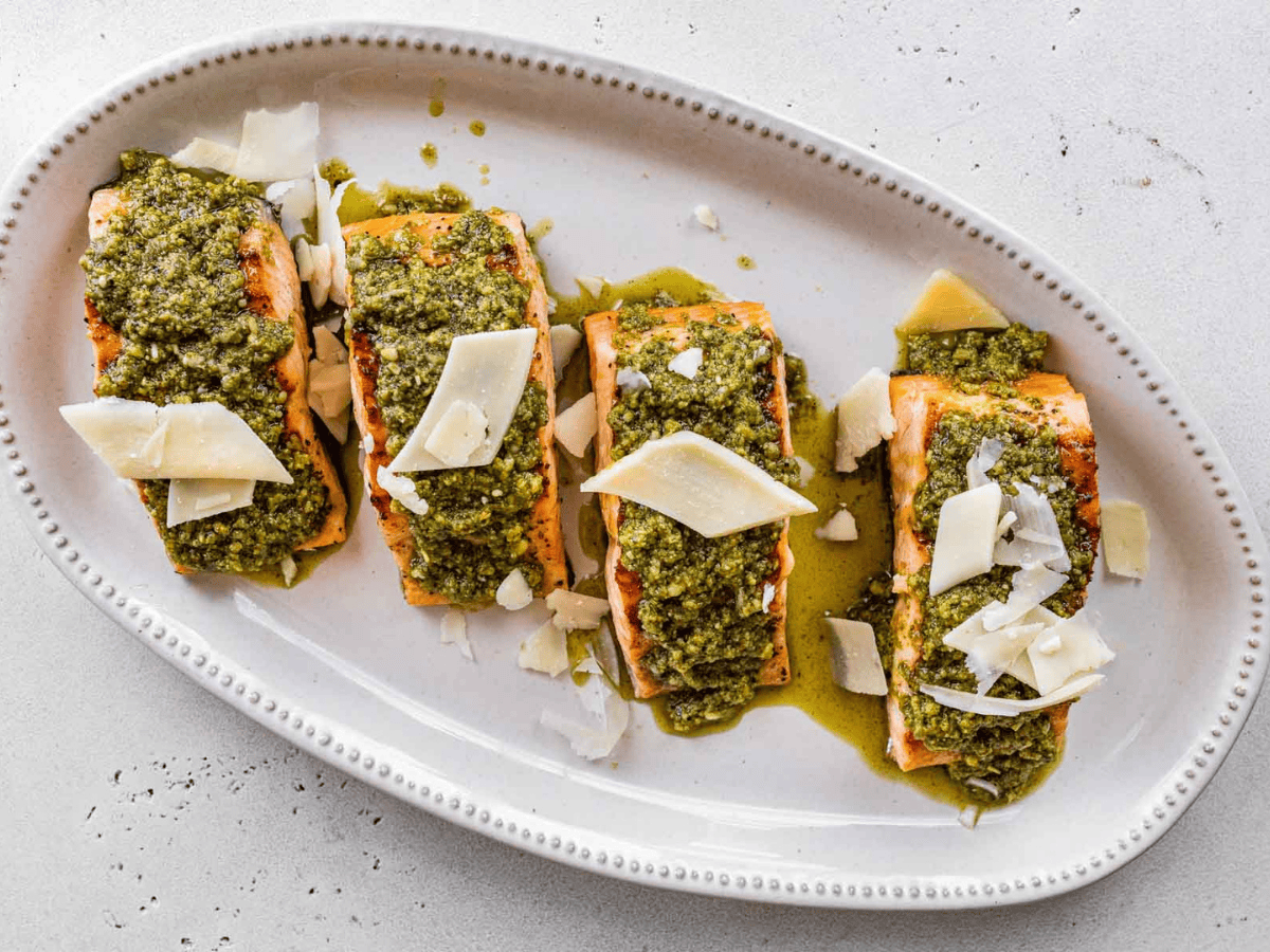 Four filets of fish plated on a white oval dish topped with a green sauce and slivers of white cheese.