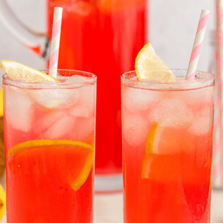 Two glasses of a rose colored drink with ice and topped with lemon wedges with a pitcher in the background.