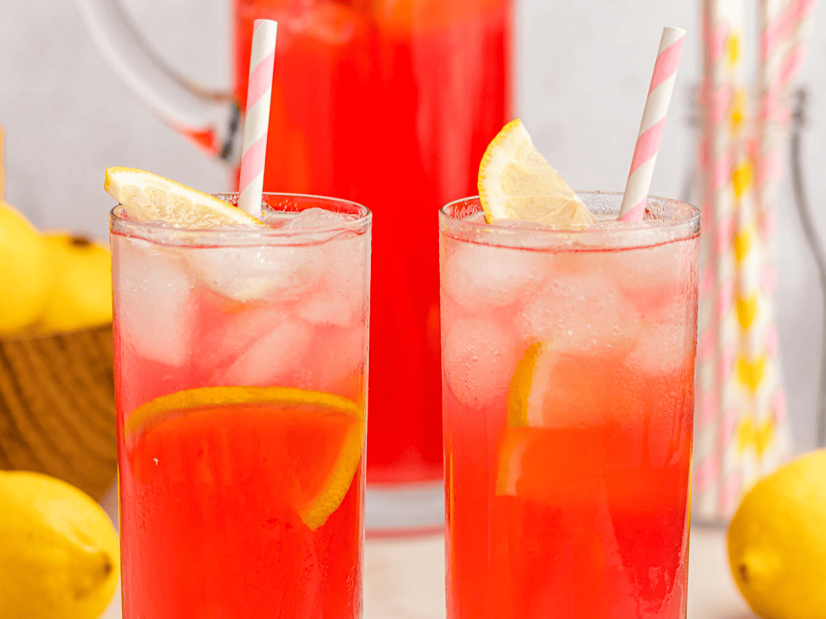 Two glasses of a rose colored drink with ice and topped with lemon wedges with a pitcher in the background.