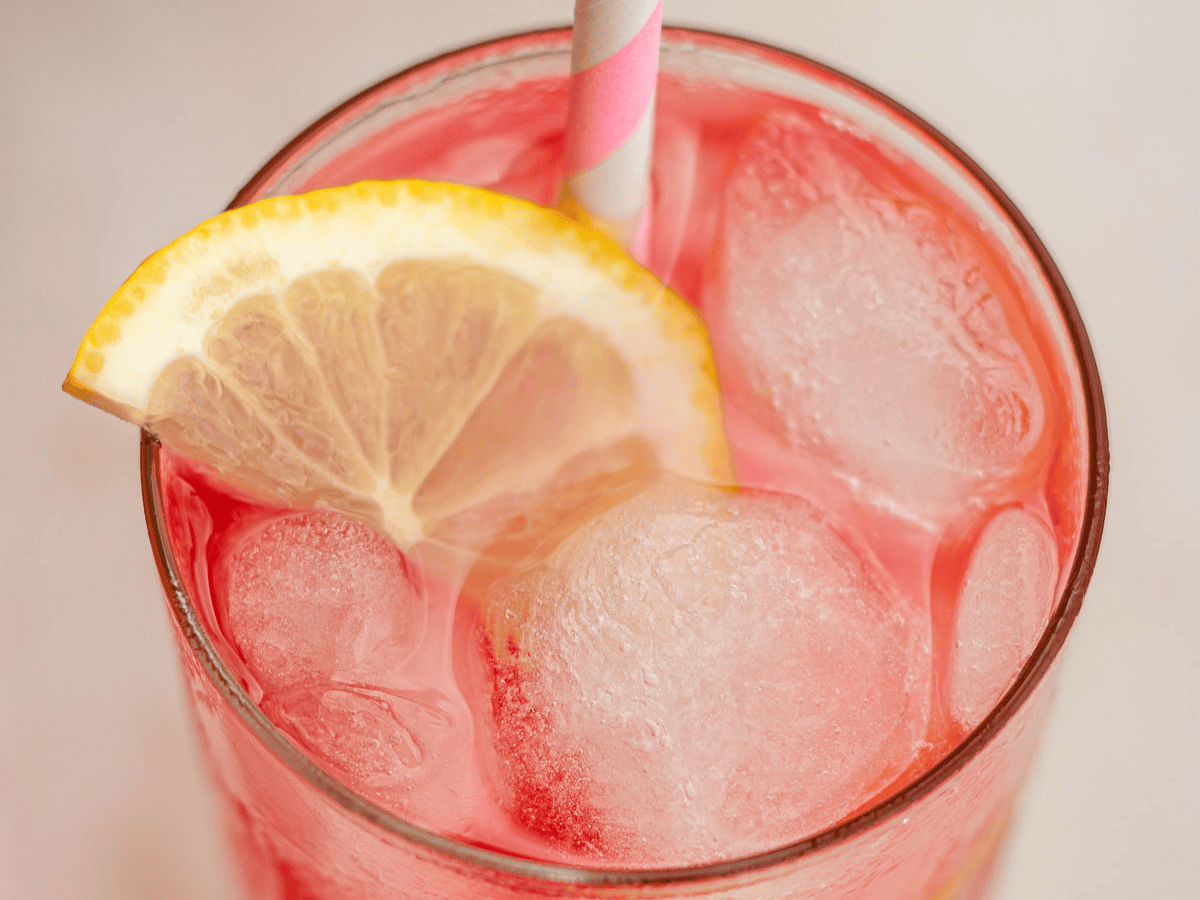Top view of rose colored drink in a glass with ice, a lemon wedge and a pink and white straw.