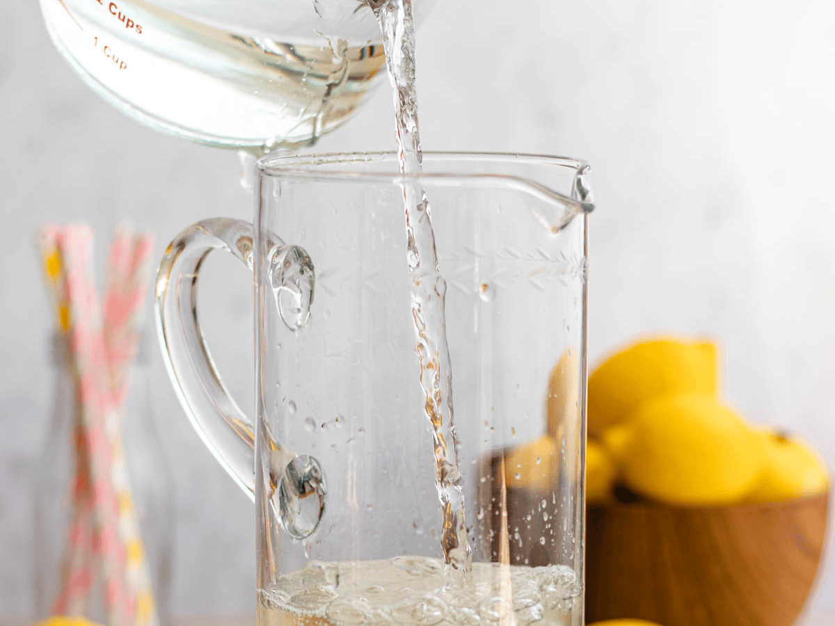 Melted sugar being poured into a glass pitcher of water with lemons in the background.