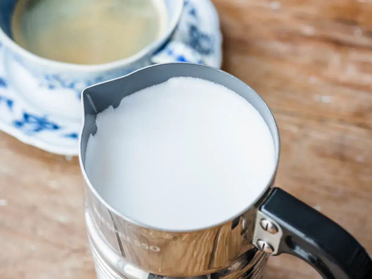 A white liquid in a silver pitcher next to a cup of hot water on top of a blue and white saucer.