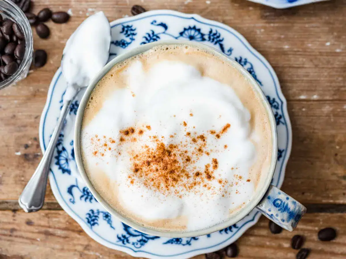 Coffee mixture with foamy top in a light blue mug on top of a white and blue saucer, with a sliver spoon next to it.