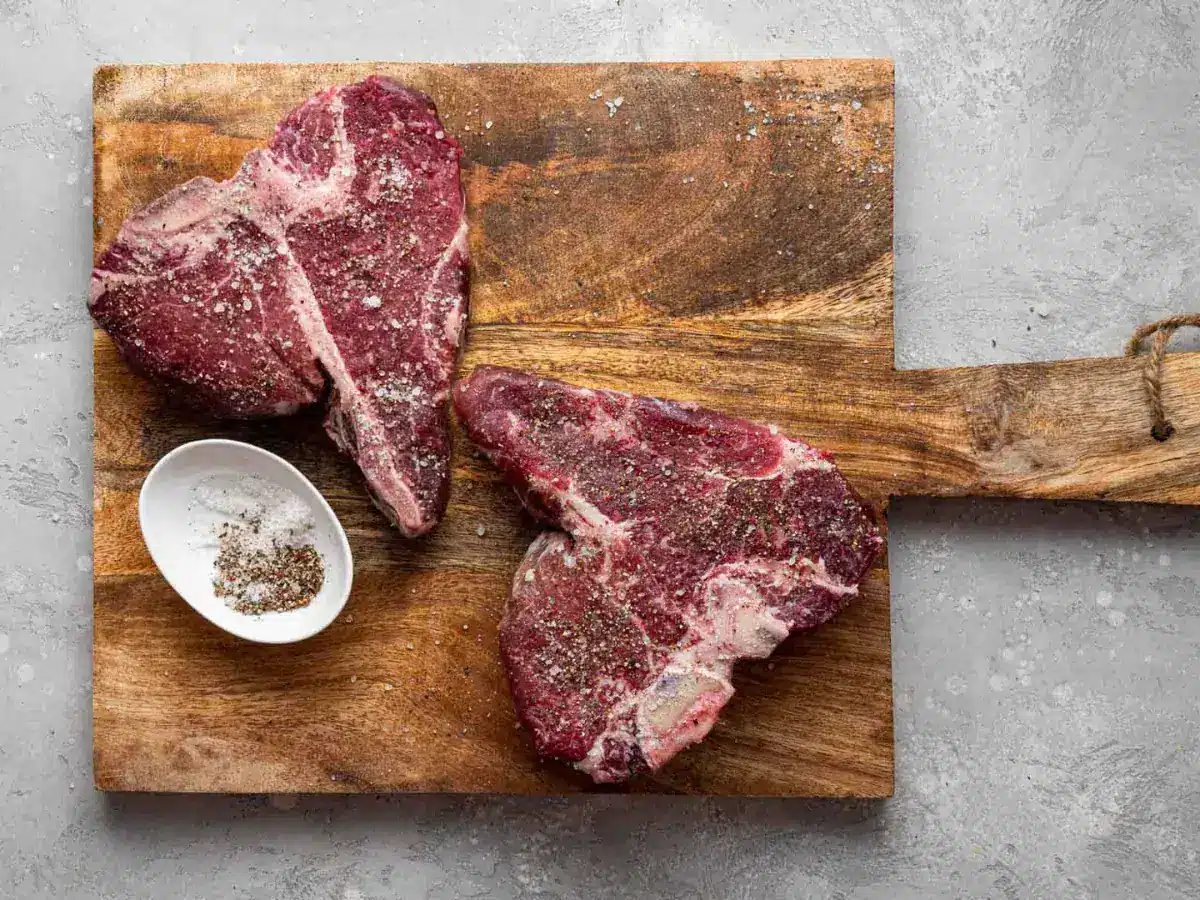 2 Steaks on wood cutting board, with a small white bowl filled with salt and pepper.