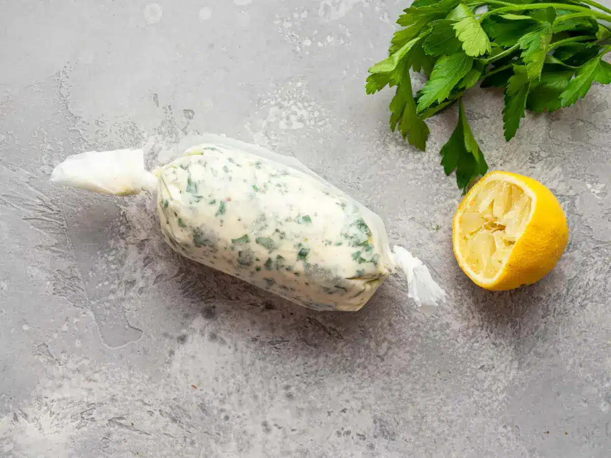 Butter mixture wrapped in a clear white paper, next to a bundle of green herbs and half a lemon.