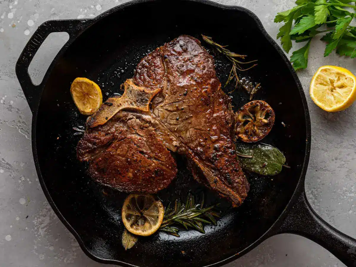 Large portion of meat with bone being cooked in a cast iron pan with lemons green herbs.