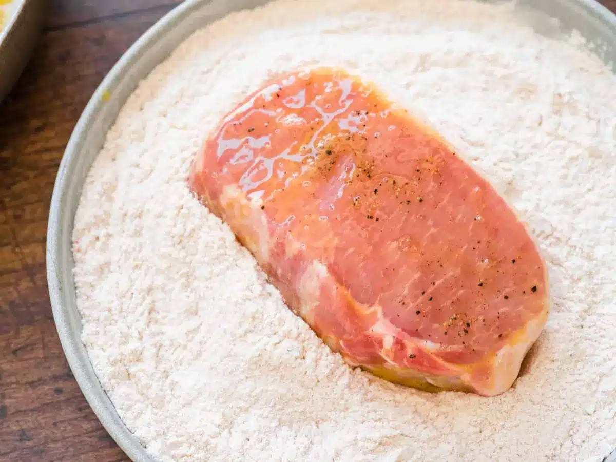 Raw pork dipped in egg mixture, sitting on seasoned flour.